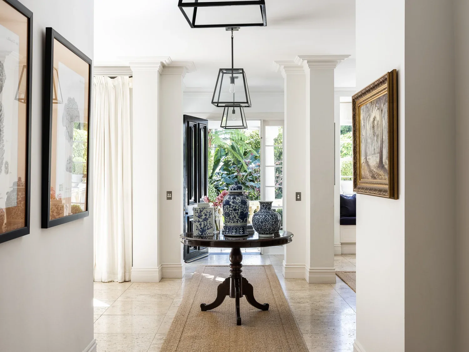 Entrance hallway with white walls, framed artwork, a round wooden table with decorative vases and flowers, and large windows with greenery outside.