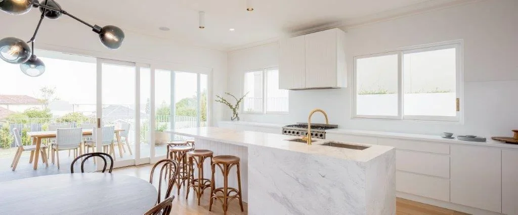 Bright, modern kitchen with white cabinets, marble island, gold faucet, and large windows.