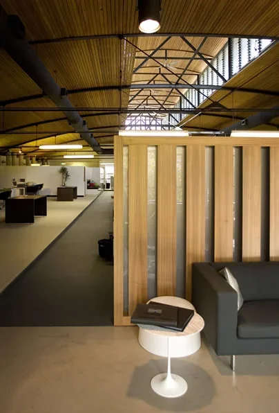 Interior of a modern office with wooden ceiling, glass walls, and open workspace. A black sofa and a small round table with books are in the foreground.
