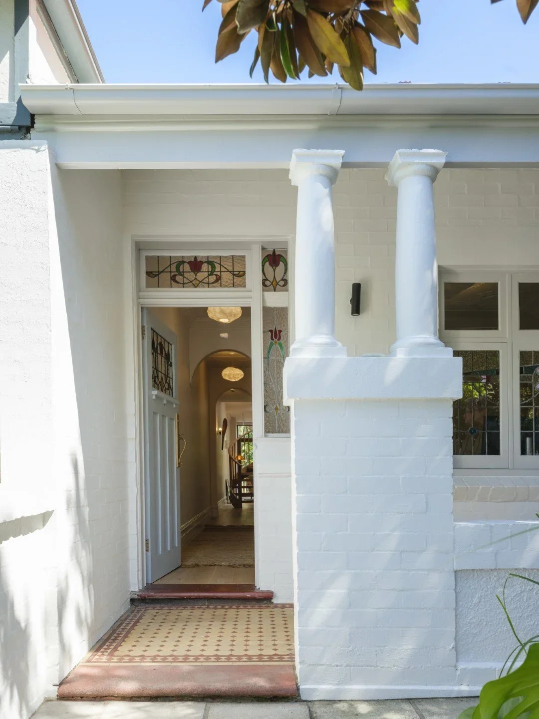 View of a white brick house entrance with a partially open door, stained glass window panels, and two white columns. Inside, a hallway with chandeliers and a staircase is visible.
