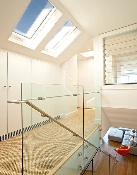 Interior view of a modern upper floor space with skylights, a glass railing, and a seating area with a gray sectional and orange chair.