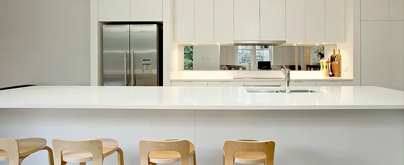 Modern kitchen with white cabinets, a large white island, and wooden bar stools. Refrigerator and a window with a view behind a mirror.