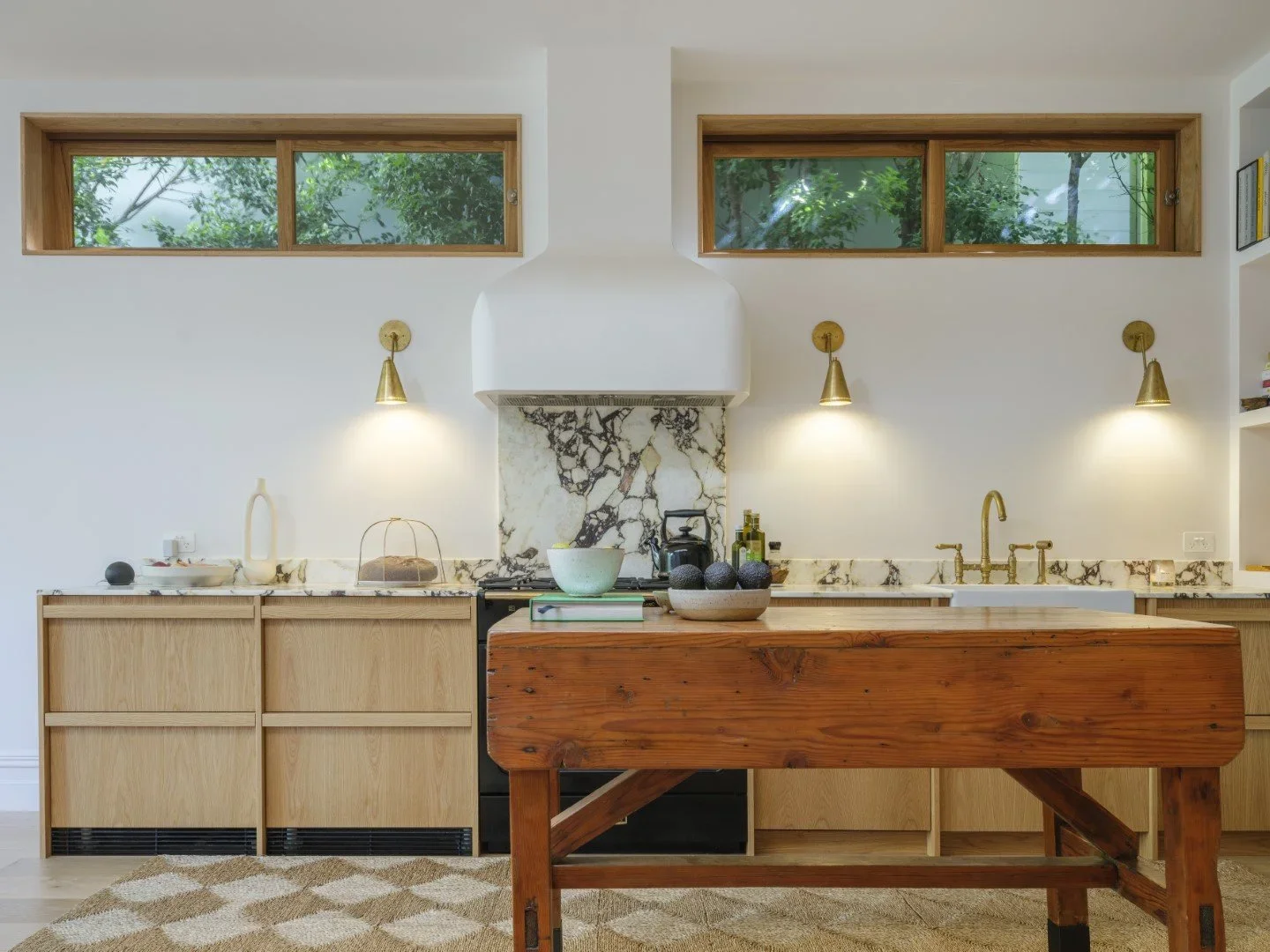 Modern kitchen with white walls, wooden cabinets, marble backsplash, gold wall sconces, and a large wooden kitchen island. Two rectangular windows above the backsplash reveal greenery outside.