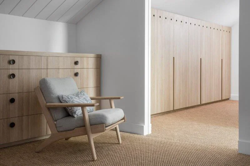 A light-colored wooden armchair with a cushioned seat and backrest, placed near built-in wooden cabinets in a minimalist room with white walls and beige carpet.
