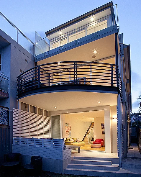Modern multi-story house with large glass windows, a curved balcony with black railing, illuminated interior, and outdoor seating area at dusk.