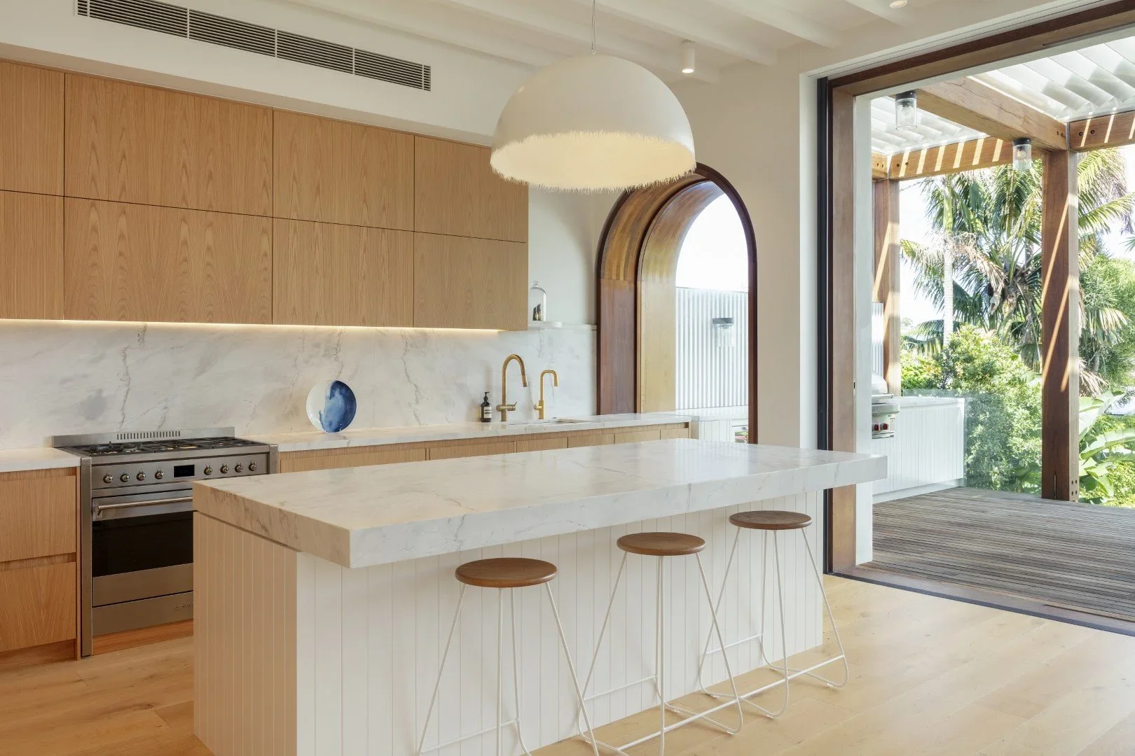 Modern kitchen with wooden cabinets, marble countertops, a white island with stools, and large sliding doors opening to a balcony with greenery outside.