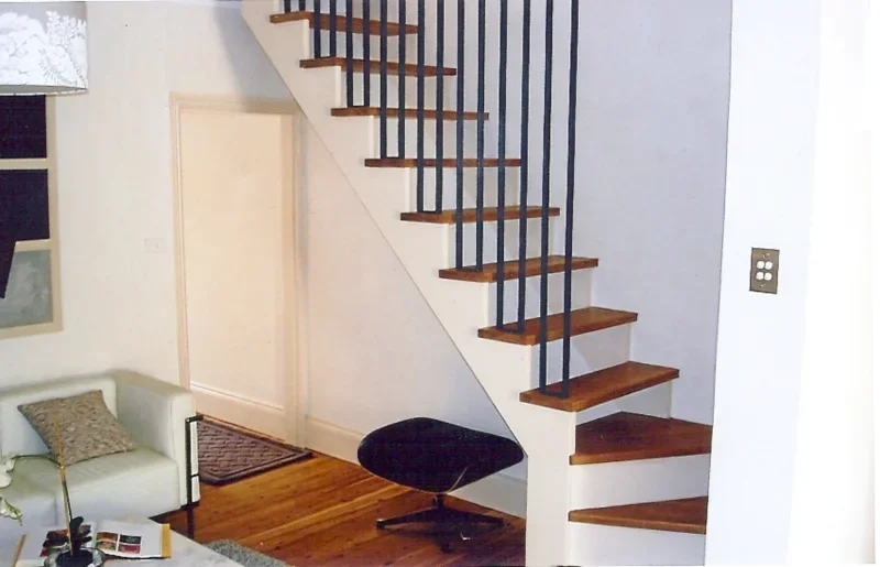 Interior of a house showing a wooden staircase with black metal railing, a black chair at the bottom, and a white sofa with a pillow in the living area.
