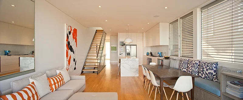 Open-concept living and dining area with a kitchen in the background, featuring a staircase, white walls, wooden flooring, a white sofa with orange and white striped pillows, a dark wooden dining table with white chairs, and large windows with blinds