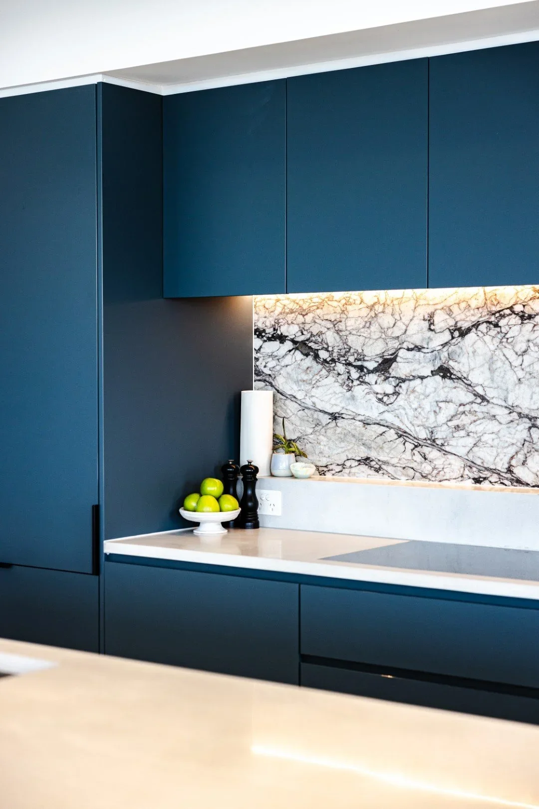 Modern kitchen with deep blue cabinets, a marble backsplash with gray and black veins, and a white countertop. A white bowl filled with green apples, and a few small kitchen items are on the counter.