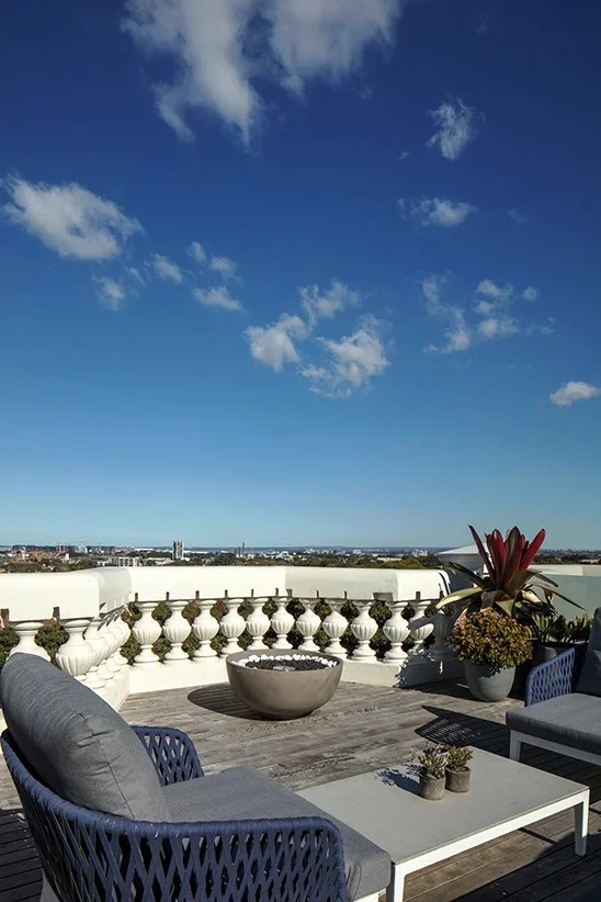 Rooftop patio with outdoor furniture, potted plants, and a city view against a blue sky with clouds.