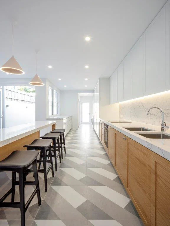 Modern kitchen with white upper cabinets, wooden lower cabinets, marble countertop, and a double sink. Bar seats with black stools along the island, pendant lights, and a windowed wall.