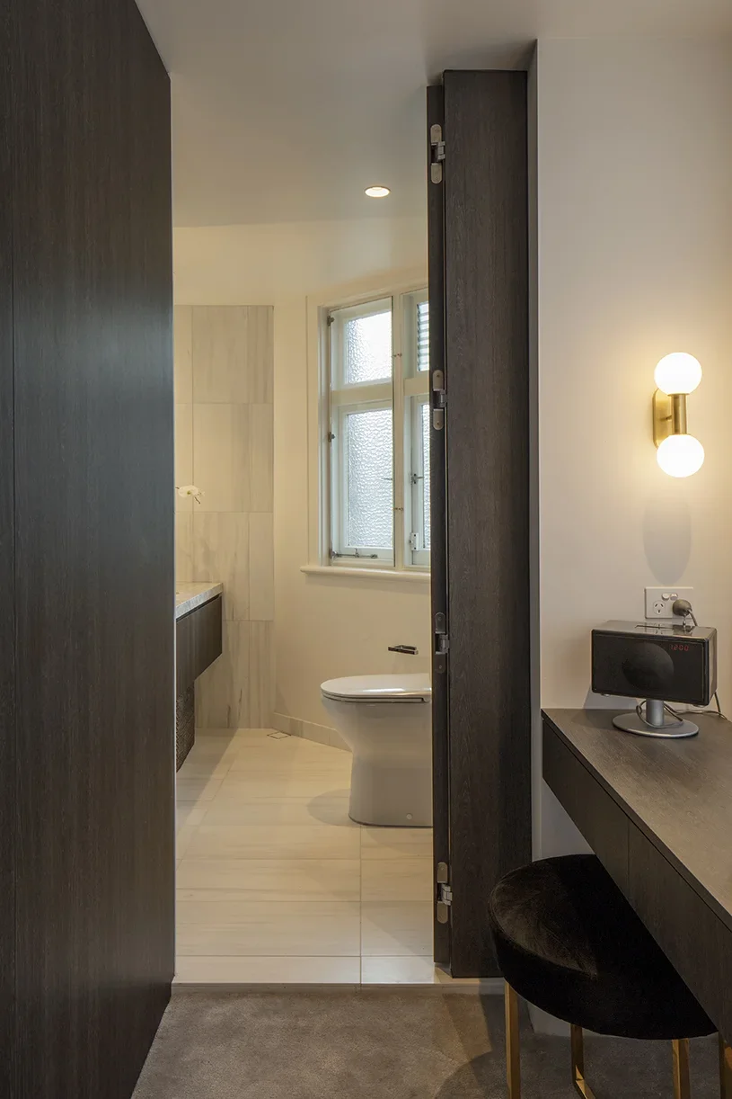 View into a modern bathroom with a toilet, frosted window, and beige tiled floor, seen from a room with a desk and a black chair.
