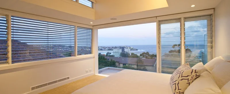 Bedroom with large windows overlooking a cityscape and water, with a white bed and decorative pillows.