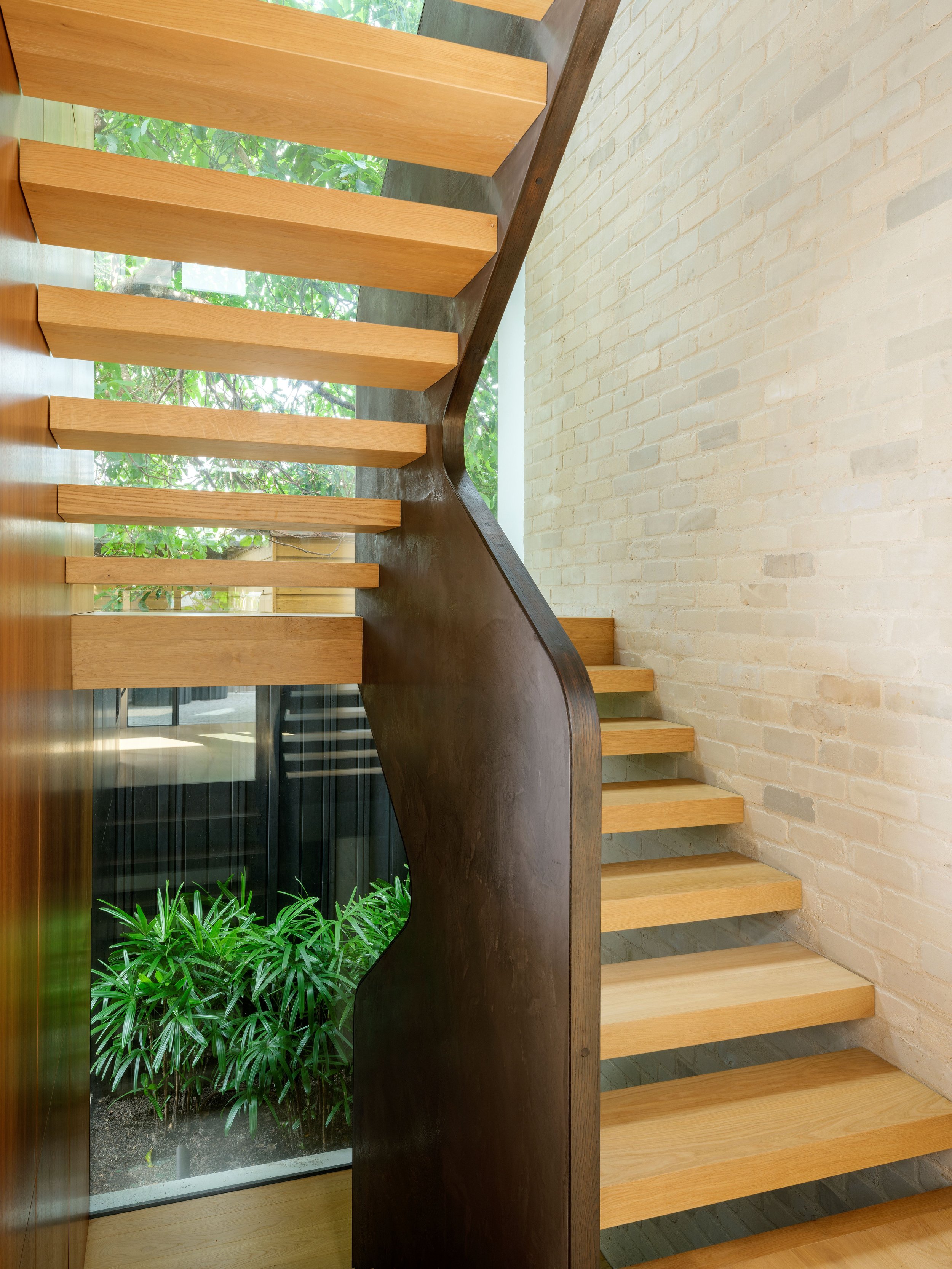 Open timber staircase at a Bronte home, with a dark balustrade, pale brickwork and a planted courtyard.