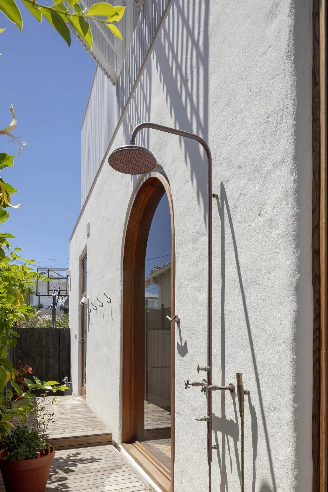 Exterior of a white house with a large arched glass door, wooden trim, and outdoor shower with a rain showerhead and exposed piping, on a sunny day with blue sky and plants nearby.