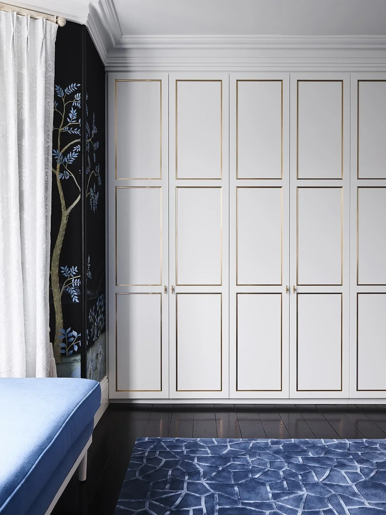 White closet with gold trim in a room with dark wooden floor and a blue patterned rug. Left side has a window with white curtains and a corner of a light blue bench.
