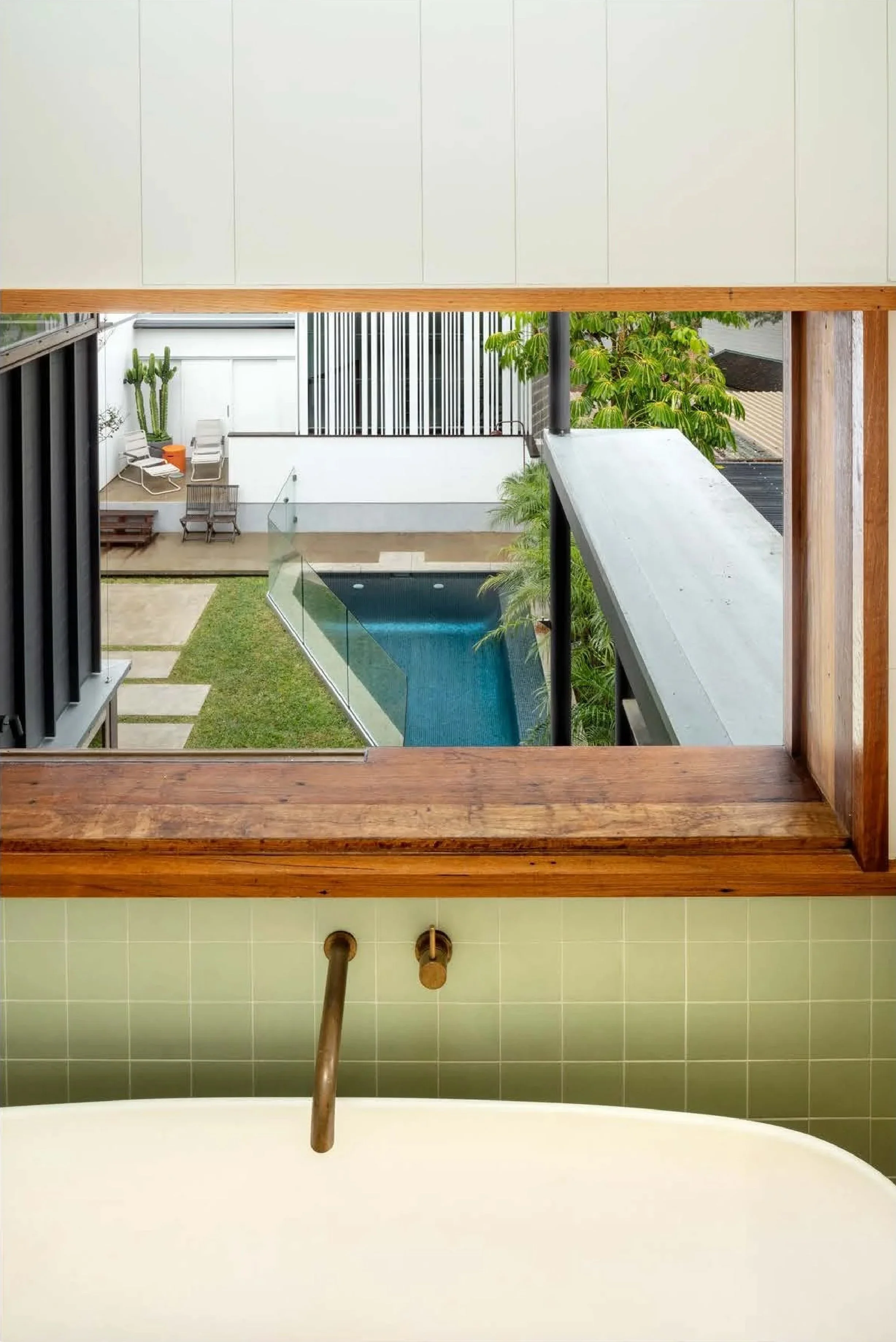 View from a bathtub looking out through a wood-framed window at a backyard with a small pool, chairs, a cactus, and greenery.