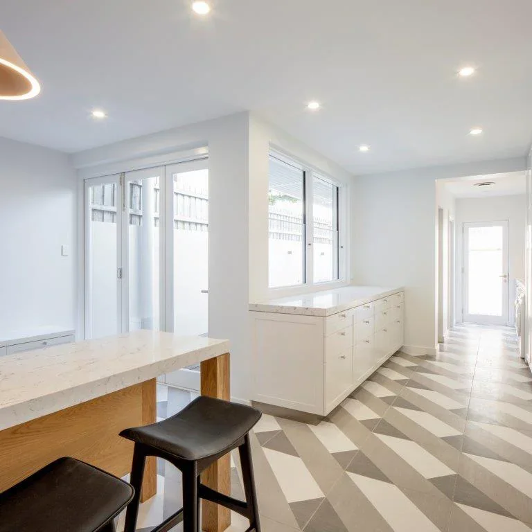 Bright kitchen with white cabinets, large windows, and a marble countertop, featuring a breakfast bar with two black stools.