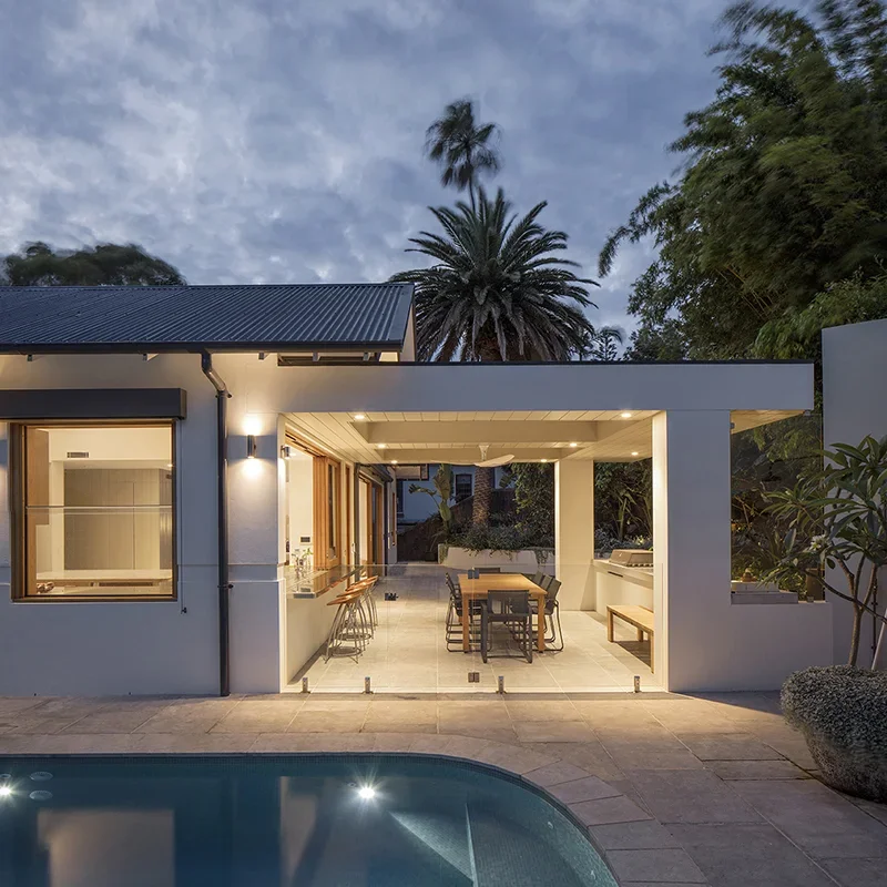 Modern backyard with swimming pool, outdoor dining area, and patio, surrounded by trees and plants during evening.