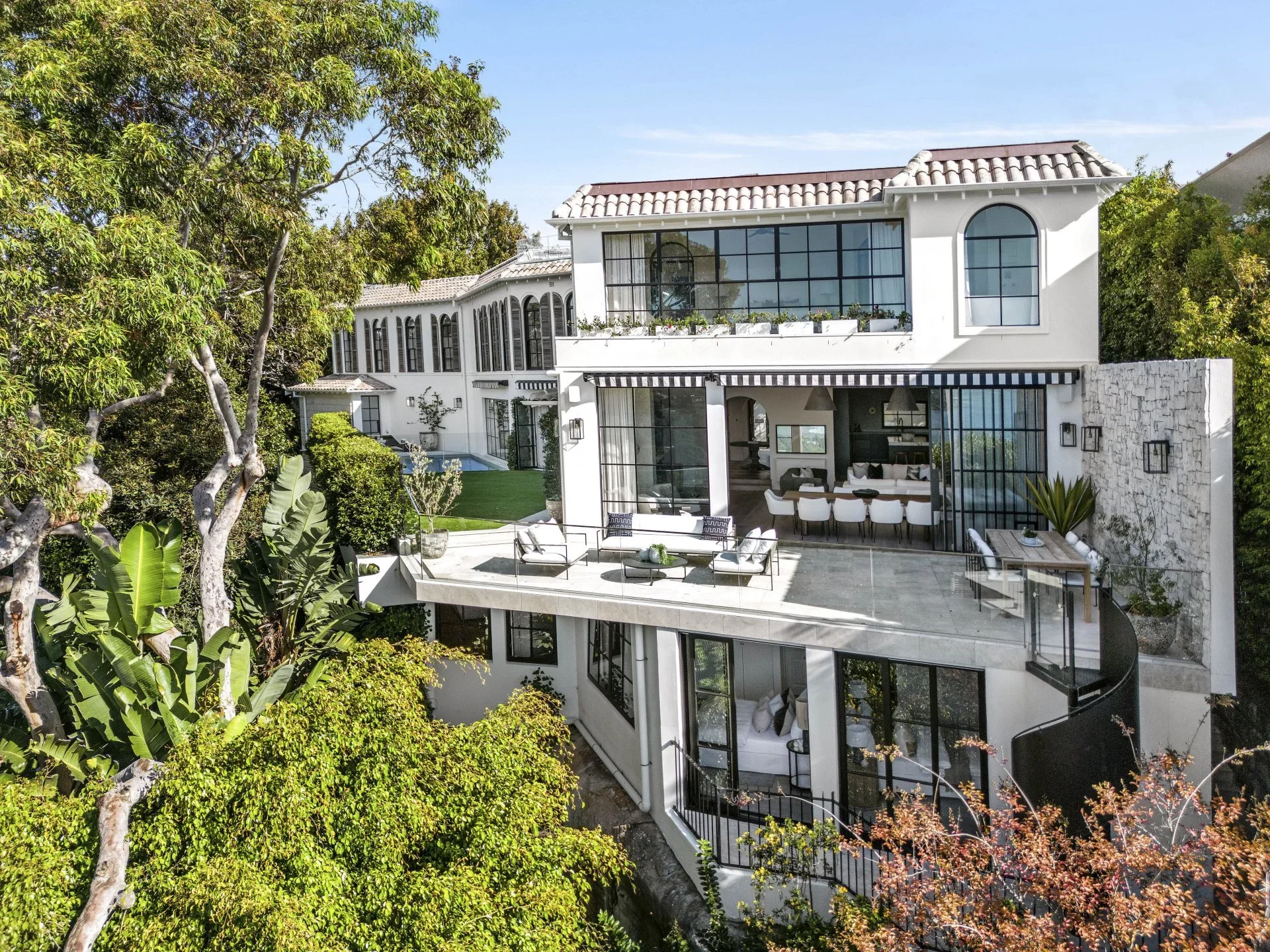 Modern multi-story white house with large glass windows and outdoor terrace, surrounded by trees and greenery.