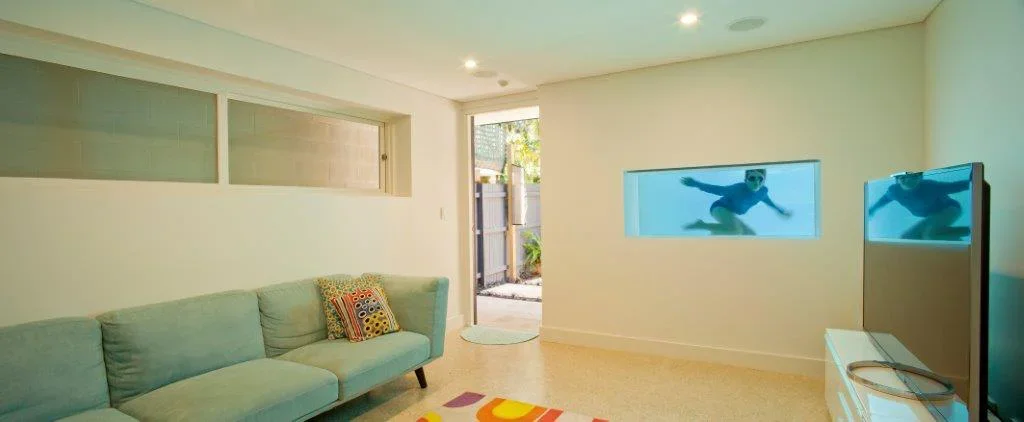 Living room with a light green sofa, colorful pillows, a colorful rug, a glass door, and a wall with a horizontal rectangular window and an aquarium with a person swimming inside.