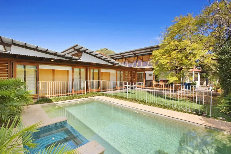 A modern backyard with a small swimming pool, surrounded by a white metal fence, with a house featuring large windows and a patio with seating, tall trees providing shade, and a clear blue sky.