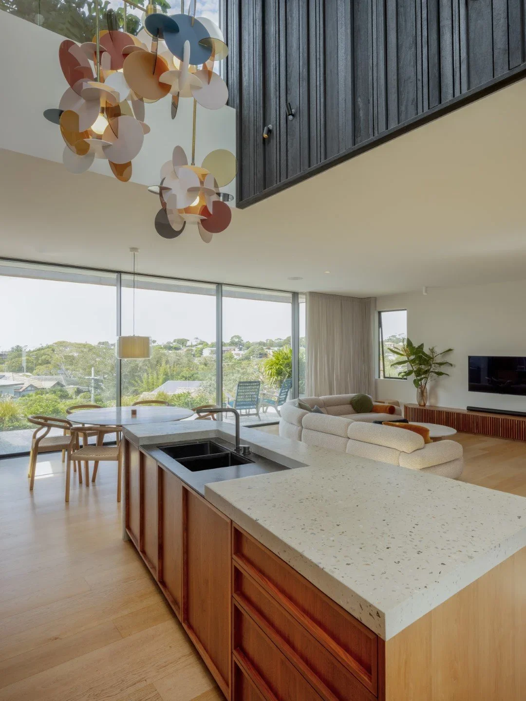 Modern open-concept living room with kitchen, large windows, and views of greenery outside, featuring a navy ceiling cabinet, a colorful pendant light, and a white sofa.