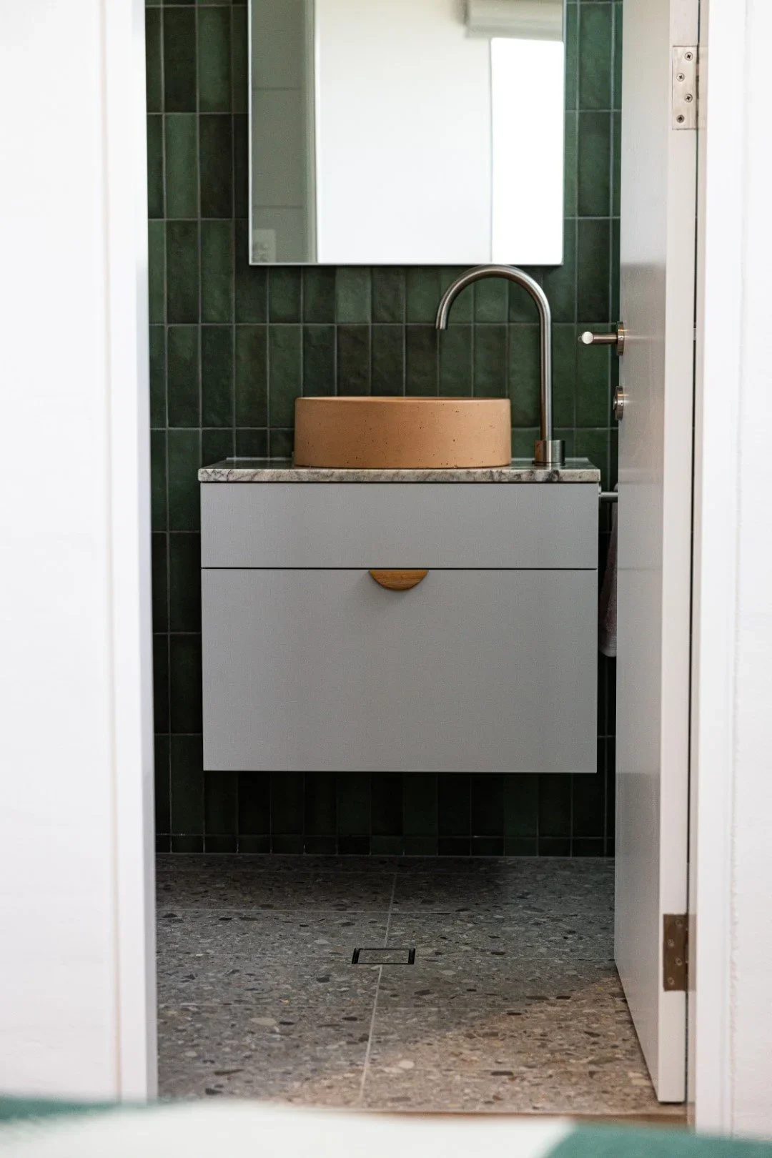 A minimalist bathroom with a white vanity, gray countertop, beige vessel sink, and green tiled wall, reflected in a mirror.