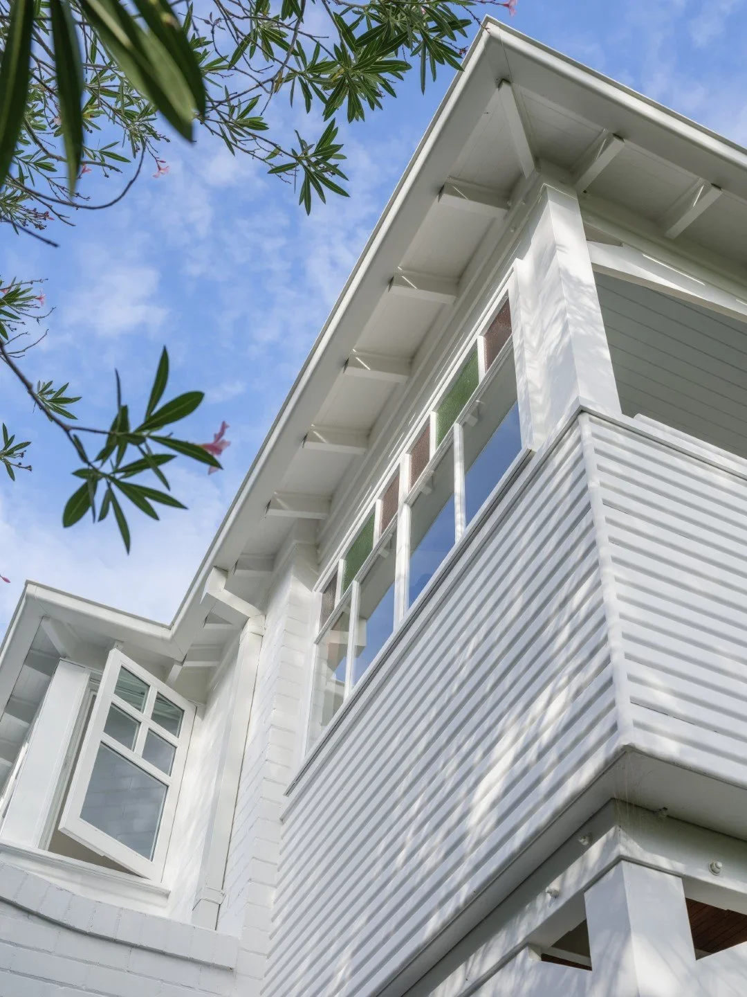 Low-angle view of a white modern house with large windows and a balcony, partly obscured by green leaves, against a blue sky with some clouds.