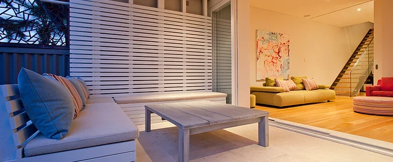 An outdoor patio area with a built-in white bench, striped pillows, and a wooden coffee table. The patio is adjacent to an indoor living room with green and pink sofas, artwork on the wall, and a staircase leading upstairs.
