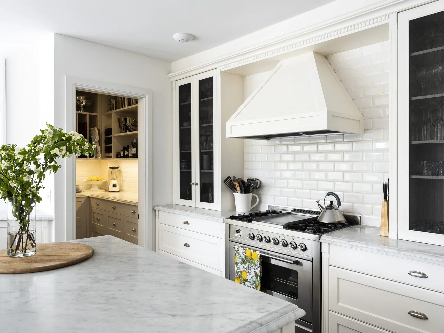 Modern white kitchen with marble countertops, a stove with a kettle, and a white range hood, with a view into a pantry area with additional cabinetry and a blender.