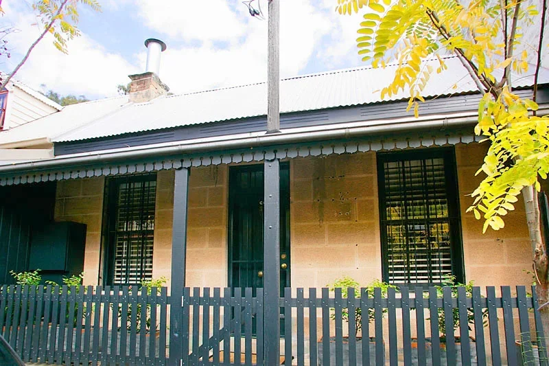 Front of a house with brick and wooden siding, metal roof, black fence, and barred windows, surrounded by trees and plants.