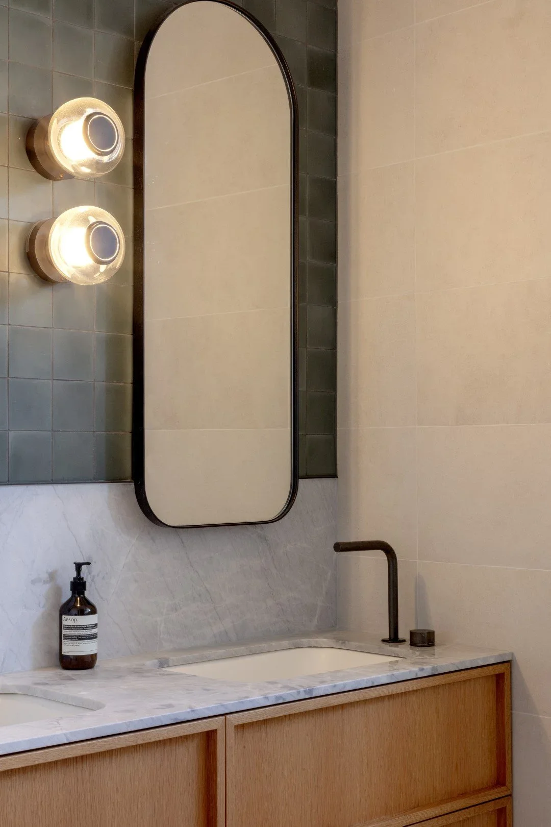 Bathroom with a marble countertop, a black faucet, a soap dispenser, an oval mirror with black frame, and two wall-mounted lights with glass covers.