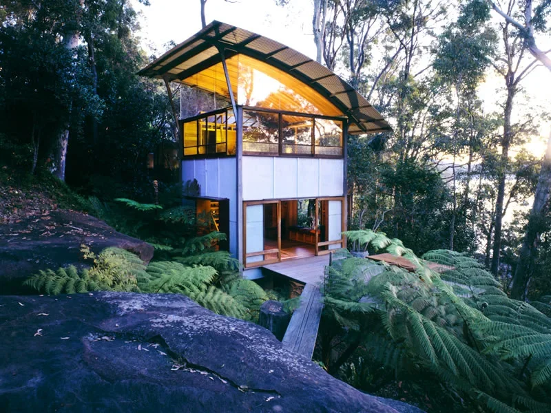 A modern house with large windows and a yellow roof, surrounded by lush greenery and situated on a hillside with a wooden walkway leading to it, in a forested area during sunset.