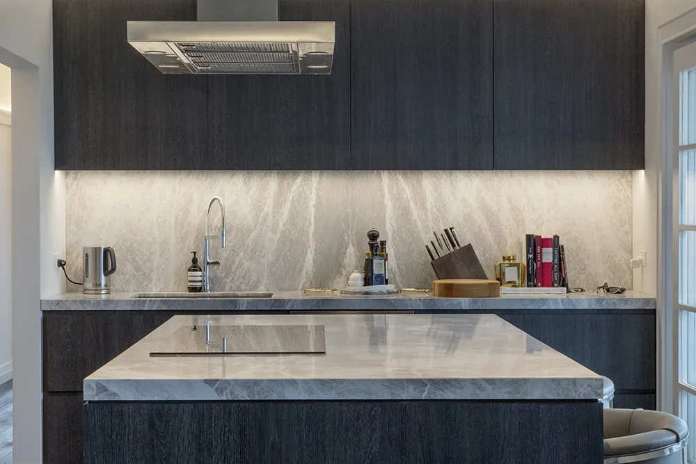 Modern kitchen with dark cabinetry, a marble countertop and backsplash, a built-in stovetop on the island, and various kitchen items including a knife block, books, and a teapot.