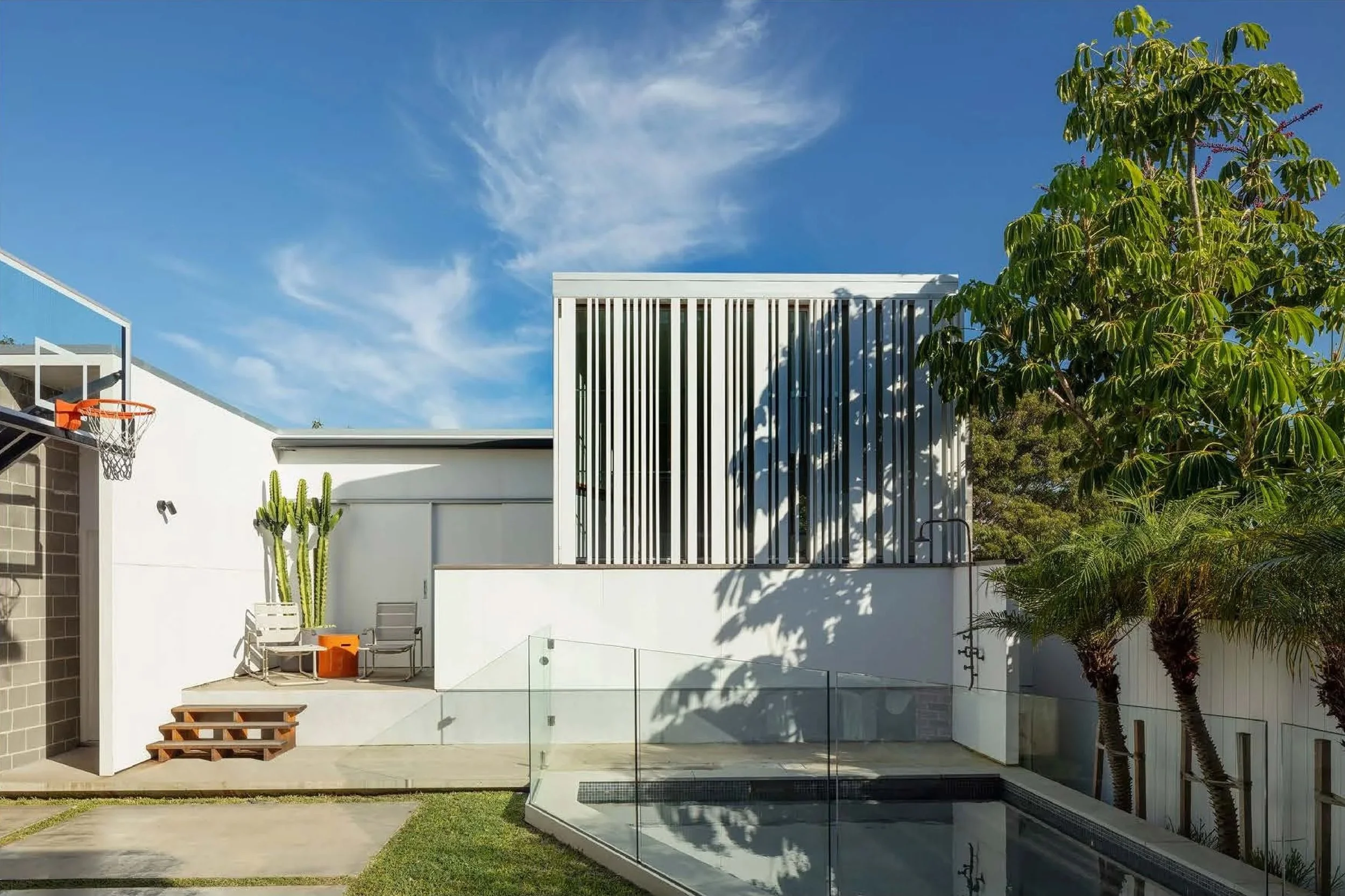 Modern backyard with swimming pool, white walls, a tall cactus, outdoor seating, a tree, and a basketball hoop under a blue sky with clouds.
