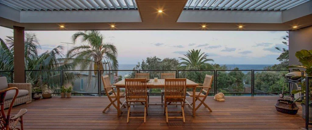 Outdoor patio with wooden dining table and chairs overlooking a lush landscape with palm trees and the ocean in the distance.