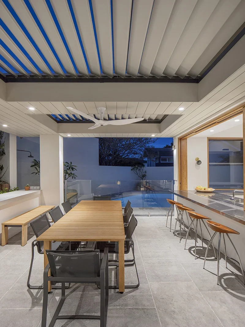 Modern outdoor patio with a wooden dining table and black chairs, barstools at a countertop, ceiling fan, and pool in the background during evening.