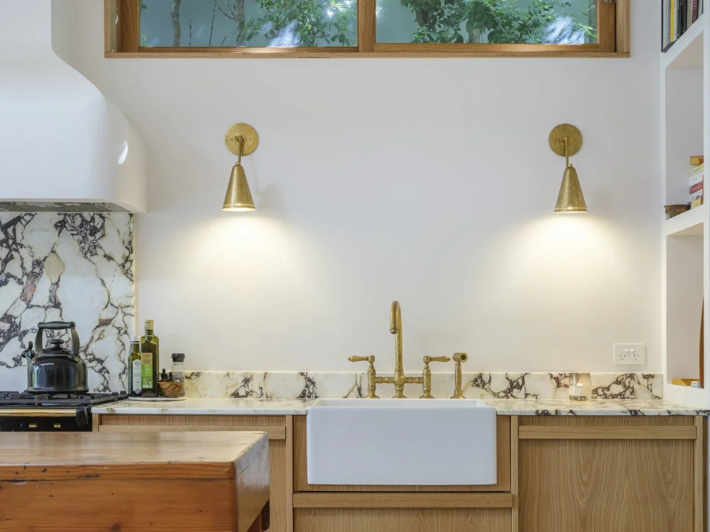 Kitchen with marble backsplash, brass faucet, wall-mounted brass lights, white farmhouse sink, wooden cabinets, and a window above.