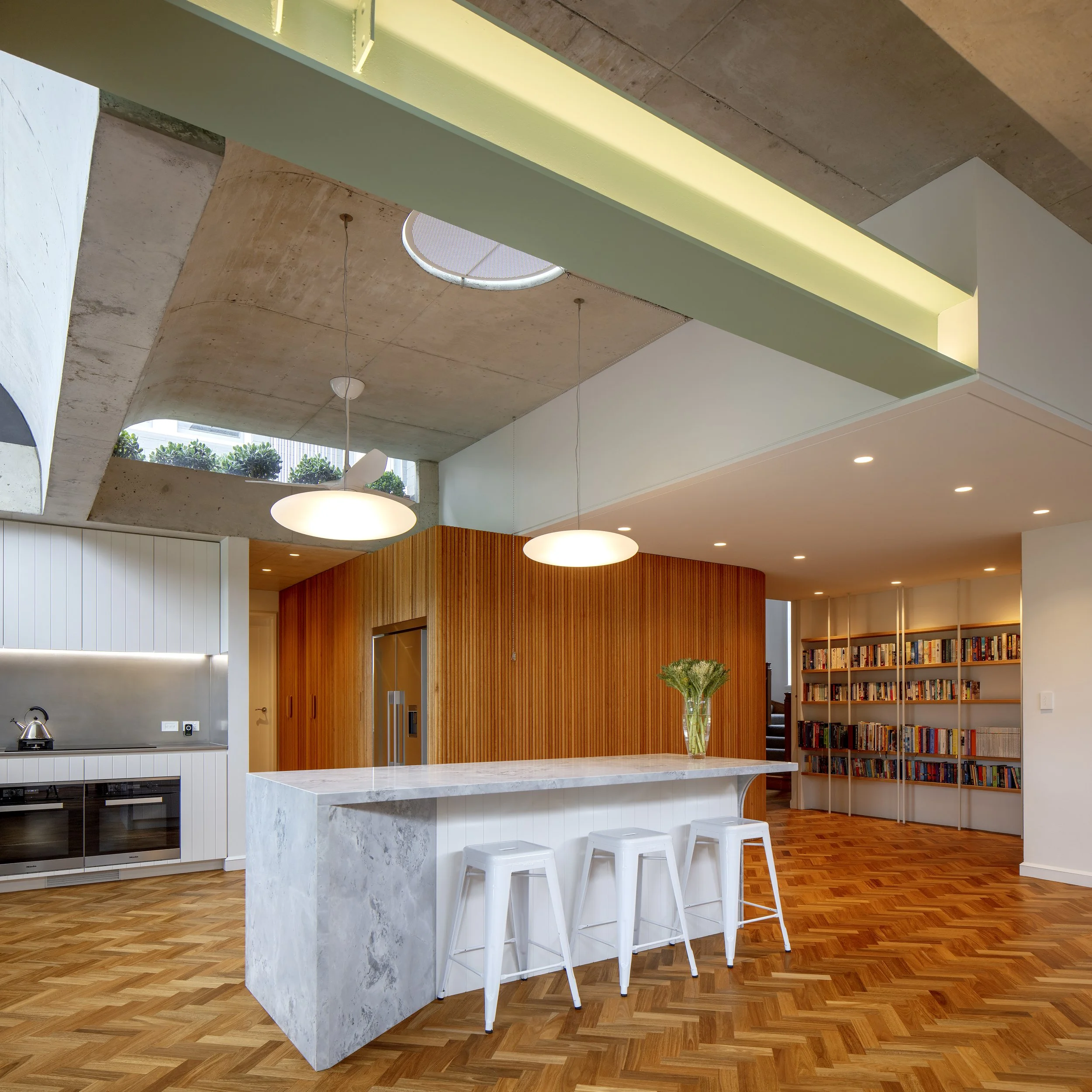 Light-filled open-plan kitchen featuring exposed concrete ceilings, circular skylights, marble island bench, curved timber joinery, and herringbone timber floors.