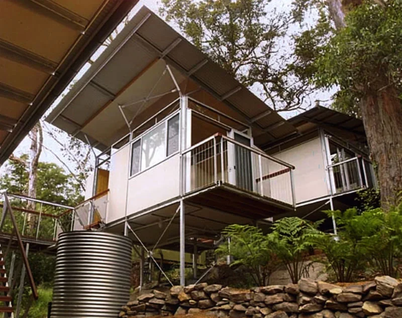 A modern tiny house elevated on stilts with a metal staircase and railing, surrounded by trees and greenery, with solar panels on the roof.