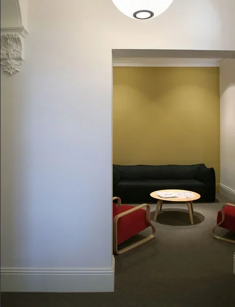A small waiting area with a black sofa, two red armchairs, and a round wooden coffee table, against a yellow accent wall in a room with white walls and decorative molding.