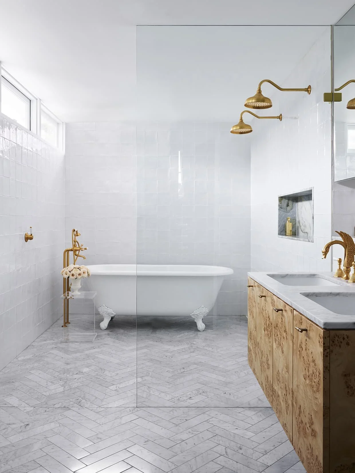 Modern bathroom with white tiled walls, a clawfoot bathtub, a wooden vanity with marble countertop, and brushed gold fixtures including showerheads and faucets.