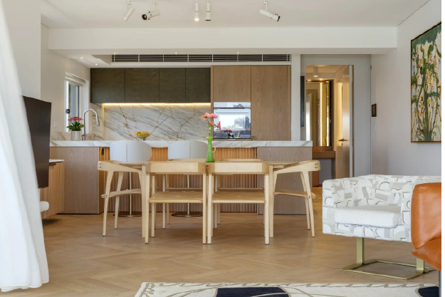 Modern open-concept kitchen and dining area with light wood finishes, marble backsplash, and a central wooden table with chairs, a white armchair, and wall art.