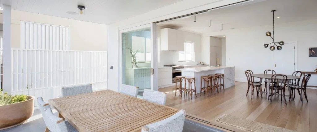 Open-concept living space featuring a dining area with a wooden table and white chairs, a kitchen with a marble island, and a separate round dining table with black chairs. Large windows and sliding glass door let in natural light.
