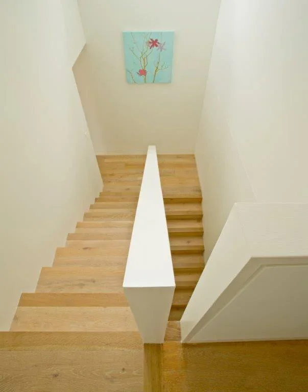 Photo of a wooden staircase seen from the top, going down, with white walls on both sides and a colorful floral painting on the wall at the bottom.