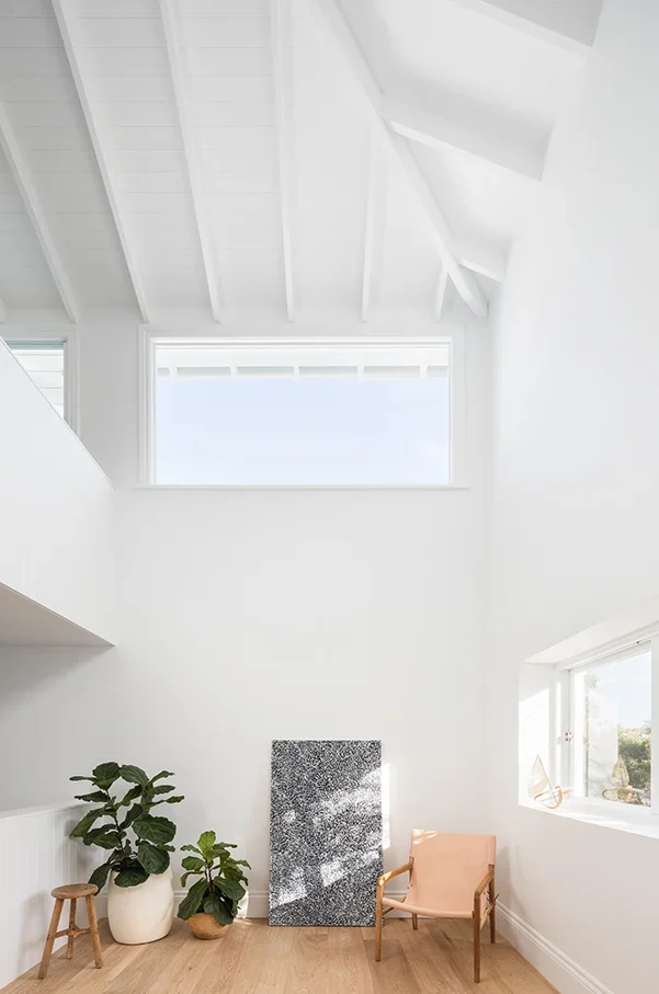 Bright white room with a skylight, two potted plants, a small wooden stool, an abstract artwork, and a peach-colored chair.