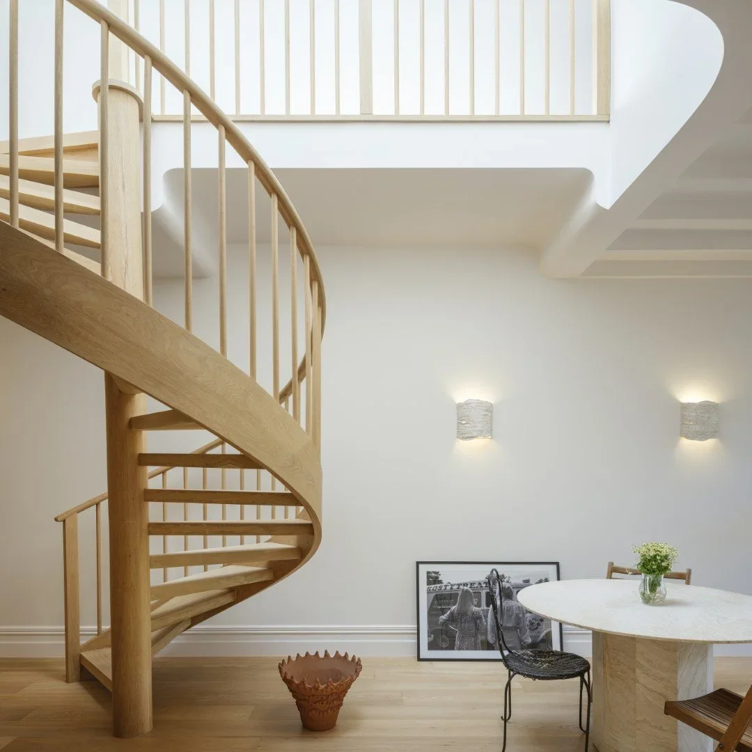Interior view of a modern home with a wooden spiral staircase, white walls, two wall-mounted lights, a framed black-and-white photo leaning against the wall, a round table with a flower vase, and a black chair.