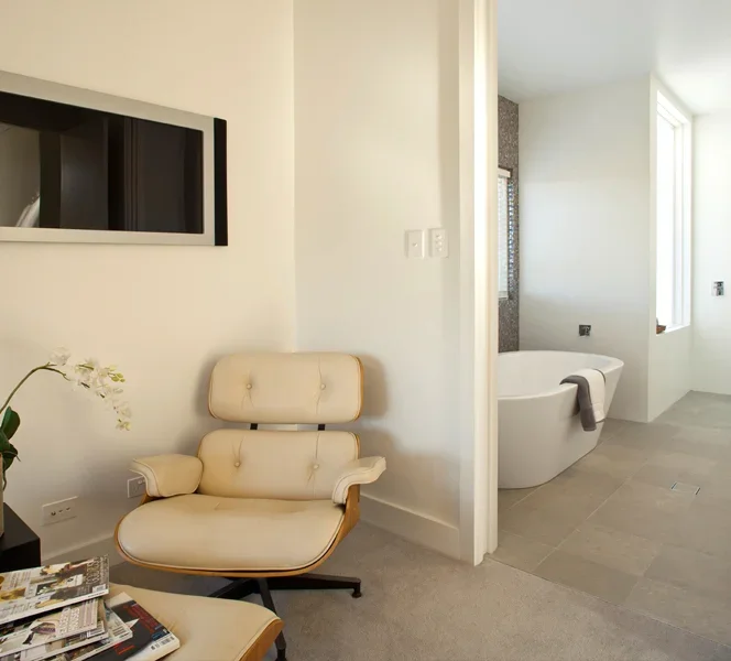A cozy living space with a white Papasan chair, a side table with magazines, and a large mirror on the wall. Adjacent is a bathroom with a freestanding bathtub and a window.