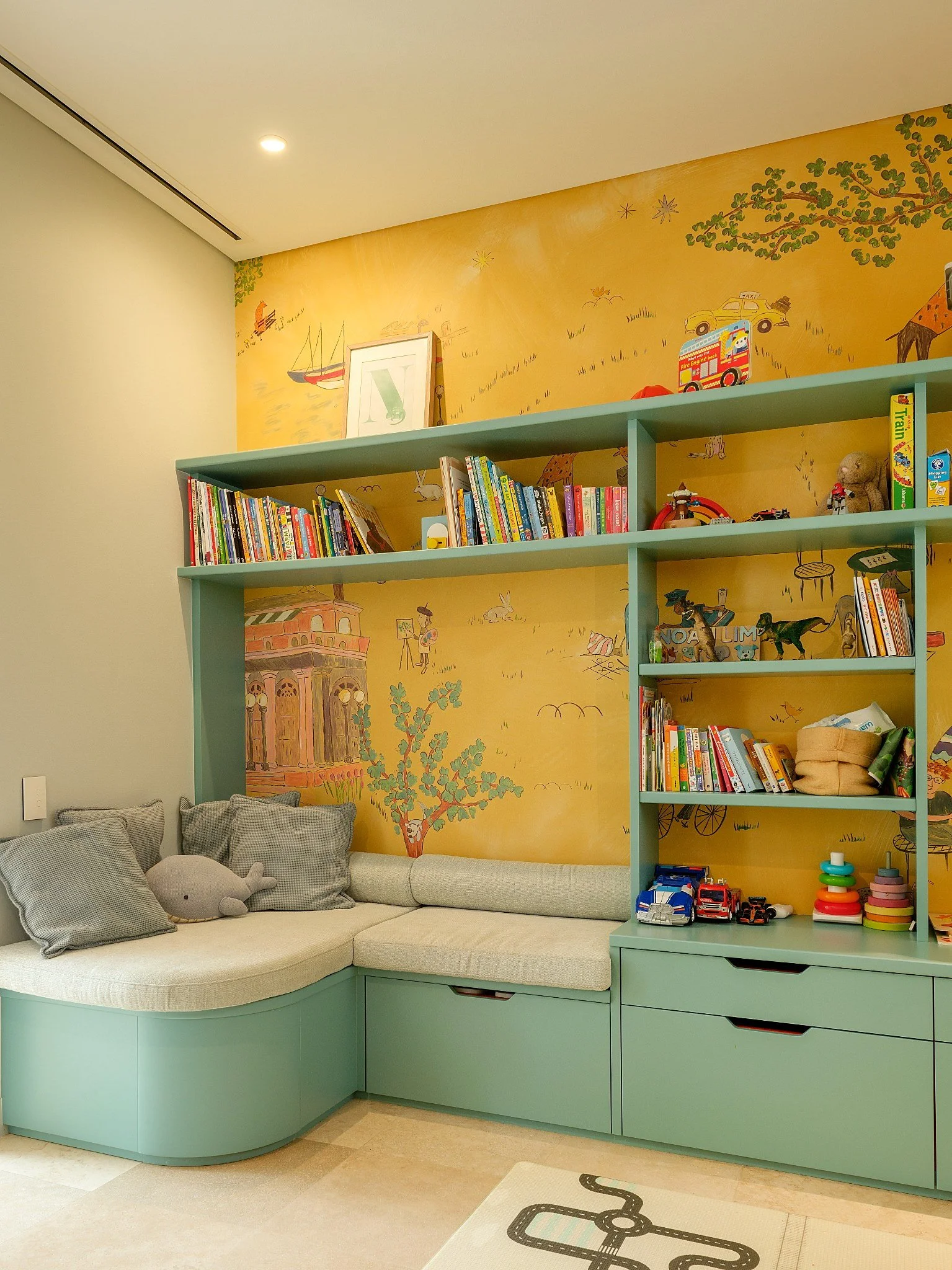 Colorful children's playroom with a mural of a park scene on the yellow wall, toy cars, ride-on toys, chairs, bookshelves, and a patterned rug.